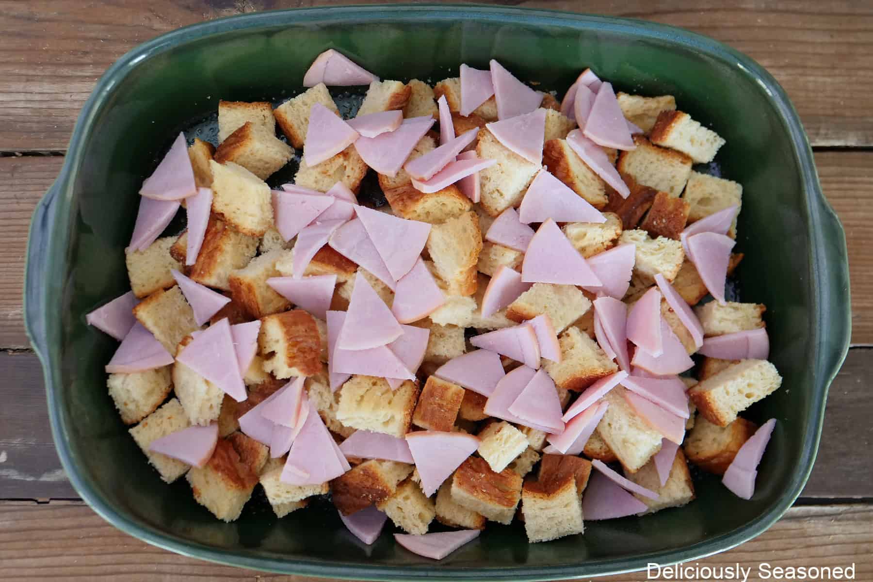 A wood surface with a green 9 by 13 baking dish filled with cubes of bread and pieces of Canadian bacon in it.