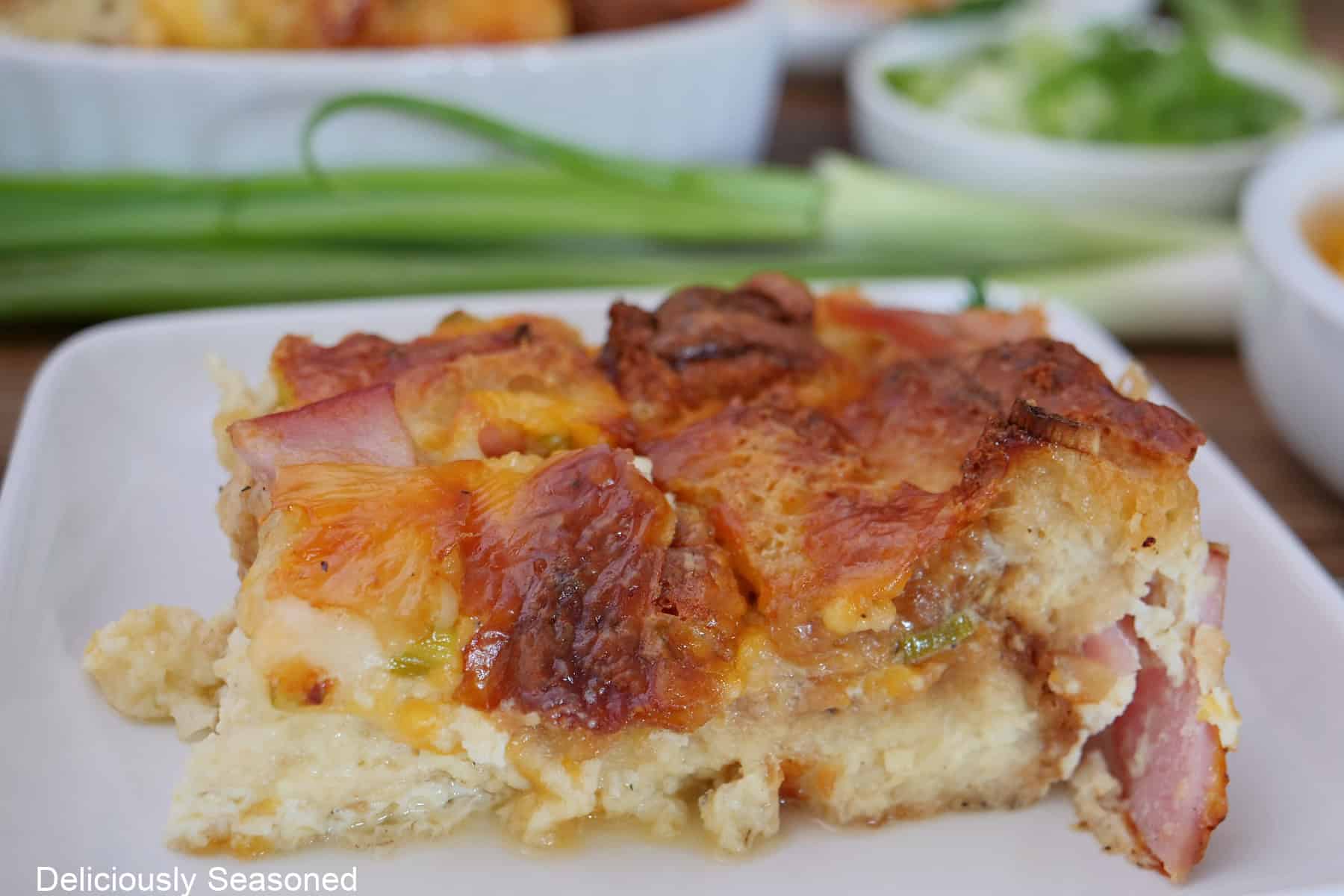 A white plate with a serving of cheesy ham bread pudding on it with green onions in the background.