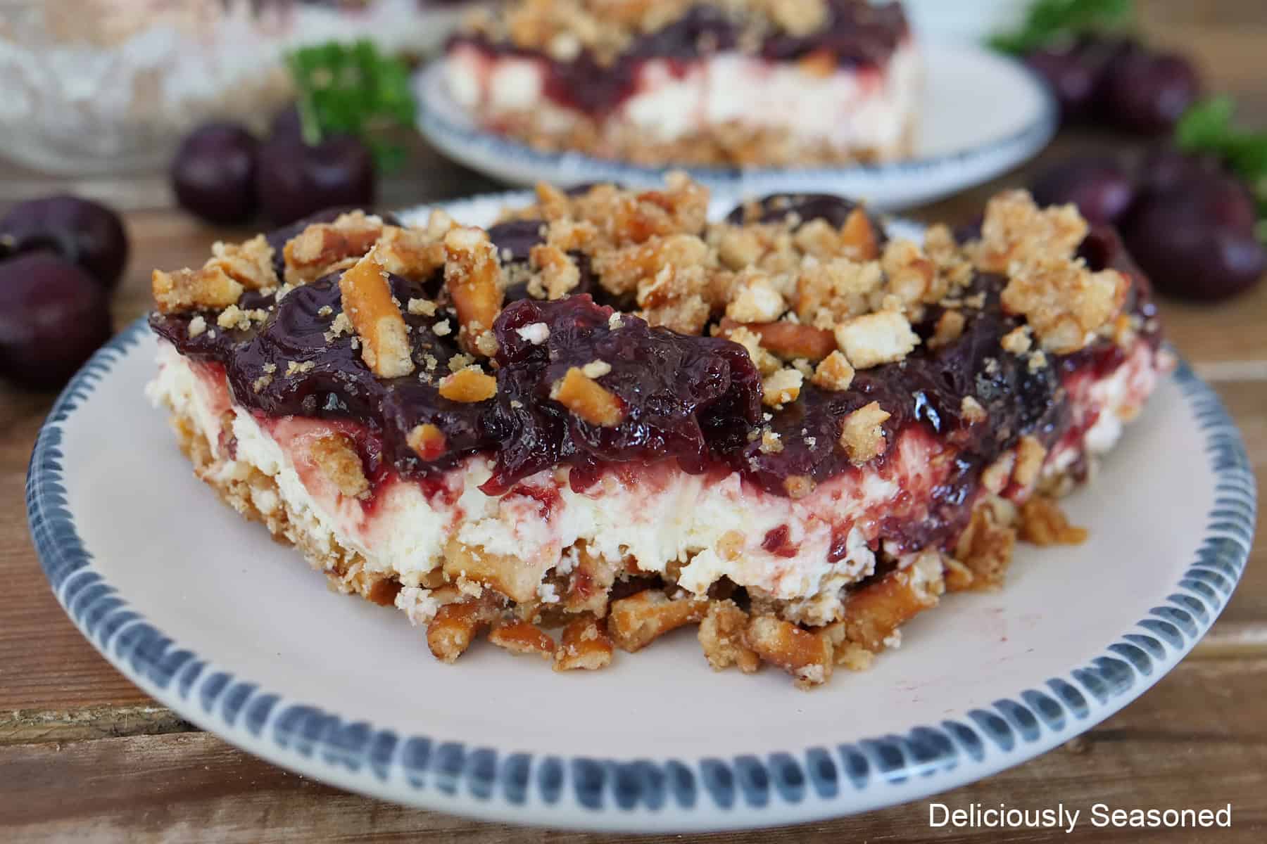 A wood surface with a white plate with blue trim with a serving of cherry pretzel salad on it with a bite taken out of it.