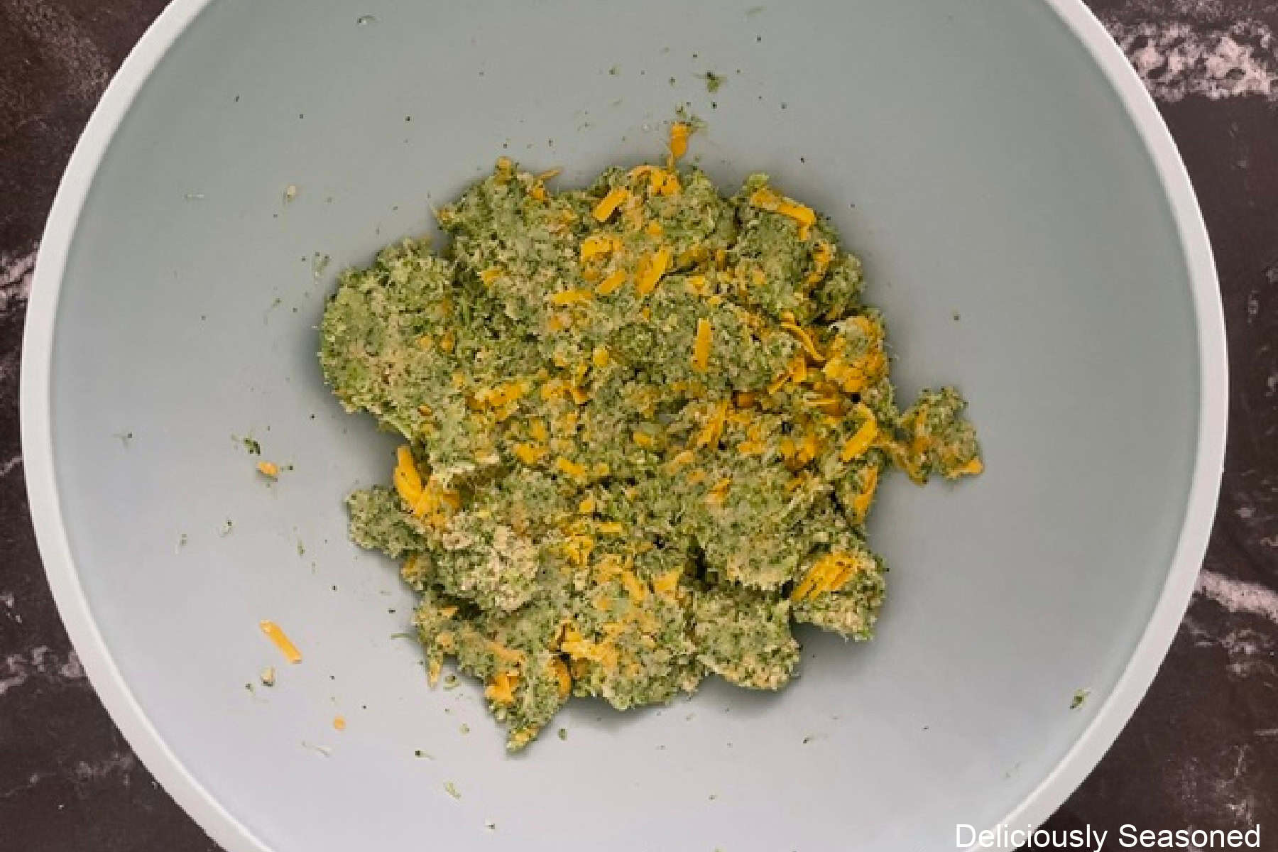 A white bowl with mashed broccoli, cheddar cheese, and a few seasonings in it all mixed together.