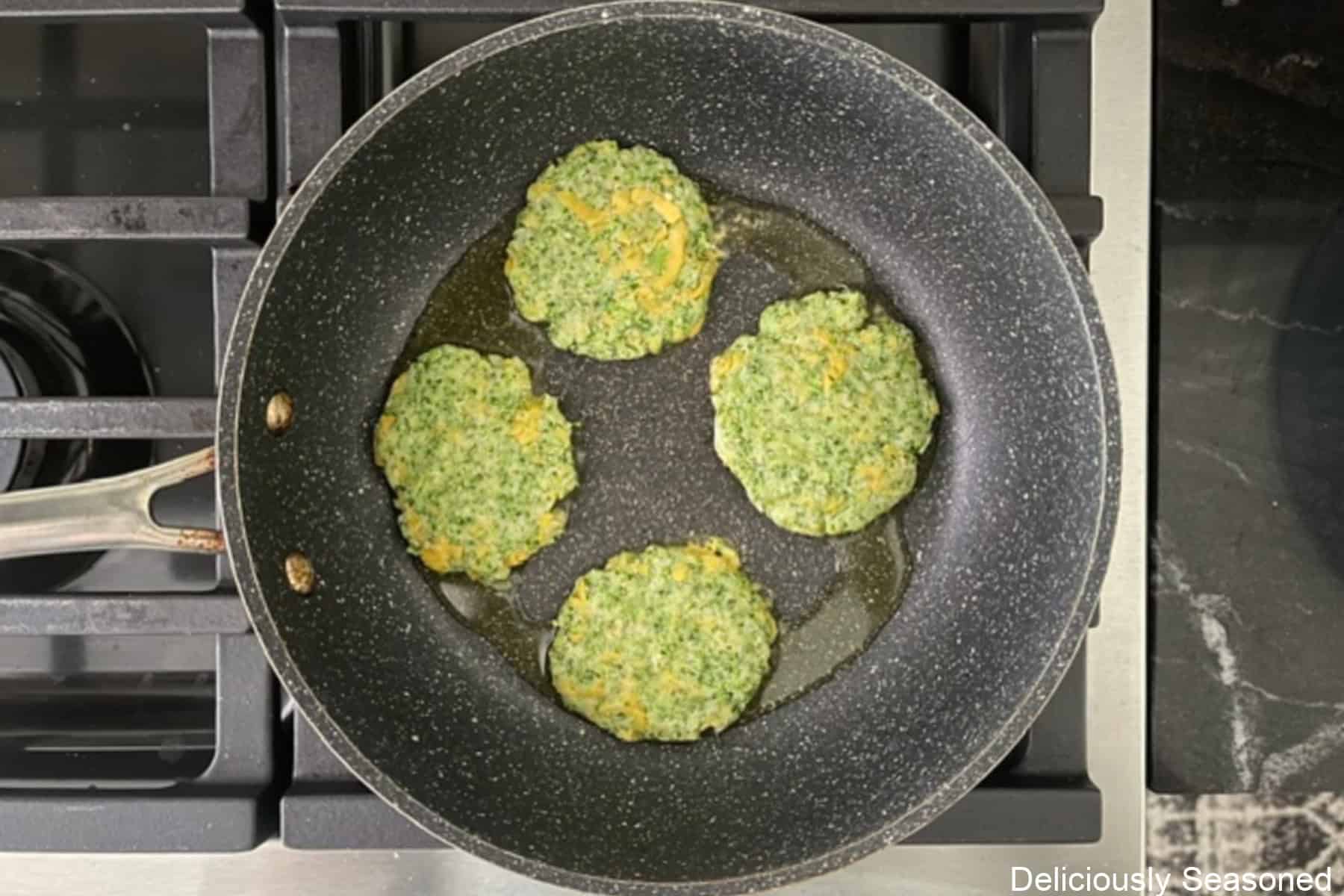 A frying pan with four broccoli cheddar patties in it frying in a little bit of oil.