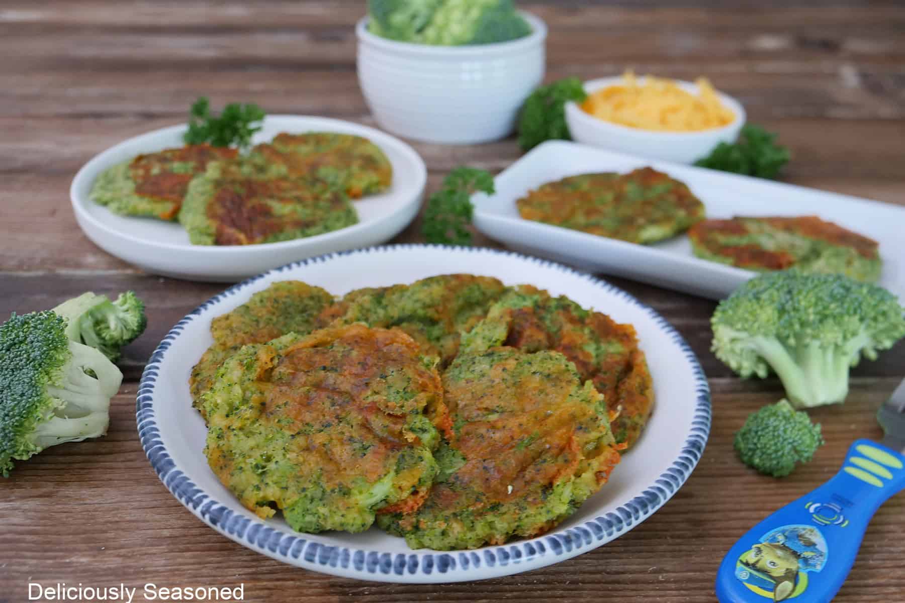 A rustic wood surface with three white plates with broccoli cheddar patties on each plate with two small white bowls filled with broccoli and cheese.