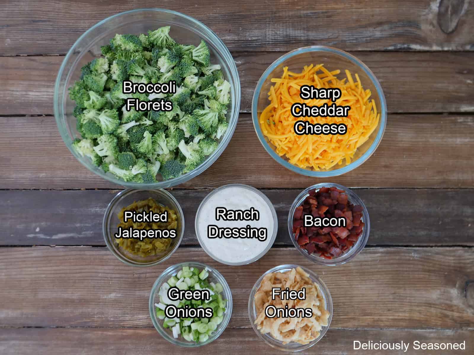 A rustic wood surface with all the ingredients placed on it to make loaded broccoli salad.