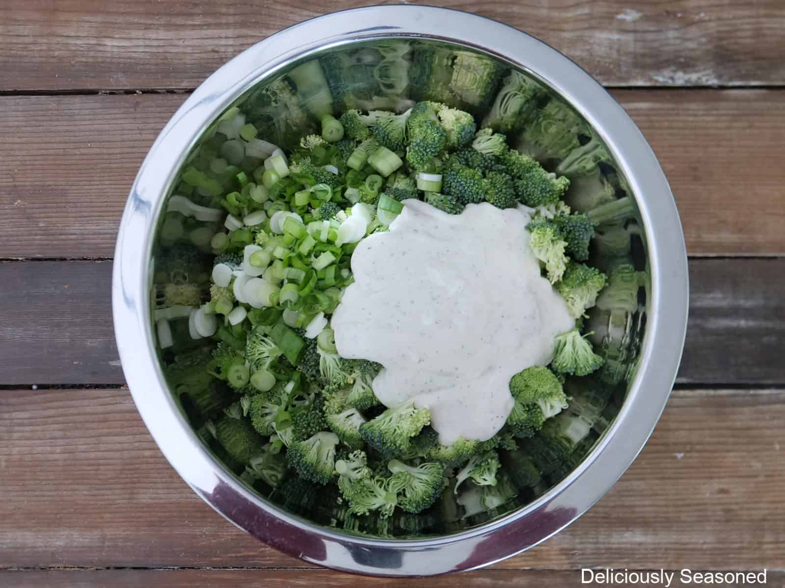 A wood surface with a silver mixing bowl filled with broccoli florets, green onions, and ranch dressing.
