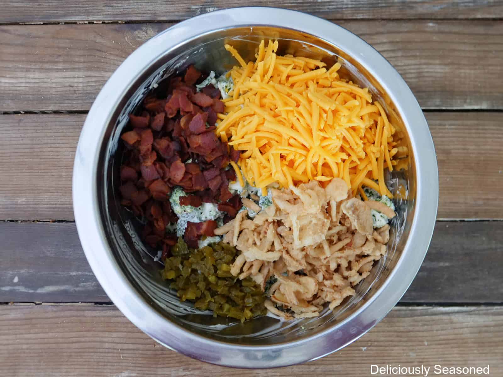 A wood surface with a sliver mixing bowl filled with crispy bacon, shredded cheese, crispy fried onions, and diced jalapenos in it.