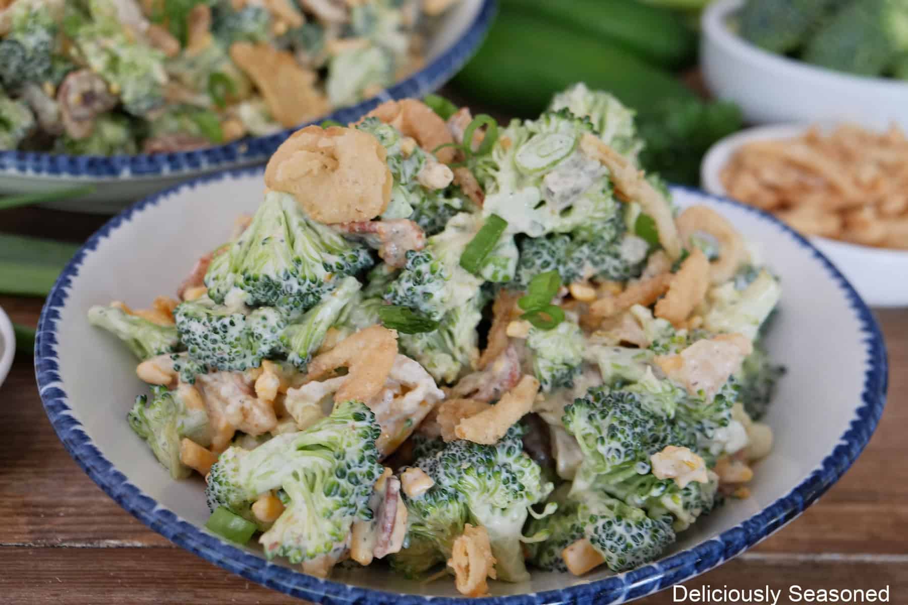 A wood surface with a blue trimmed white bowl filled with a serving of loaded broccoli salad.