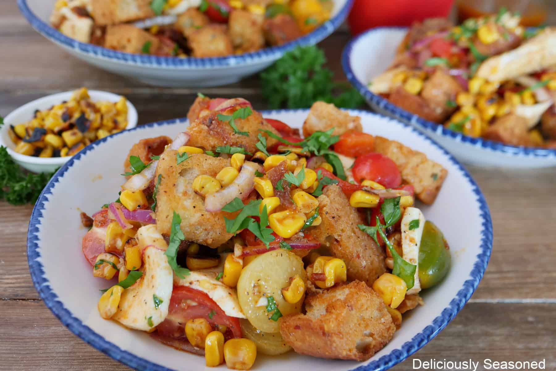 A white bowl with blue trim filled with roasted corn panzanella salad in it with two additional bowls in the background.