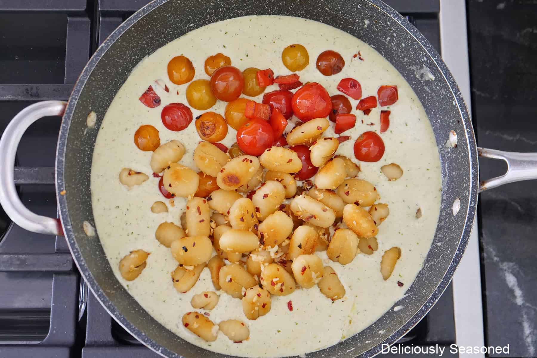A frying pan on the stove with a creamy alfredo sauce with potato gnocchi and cherry tomatoes and red bell pepper added before being combined.
