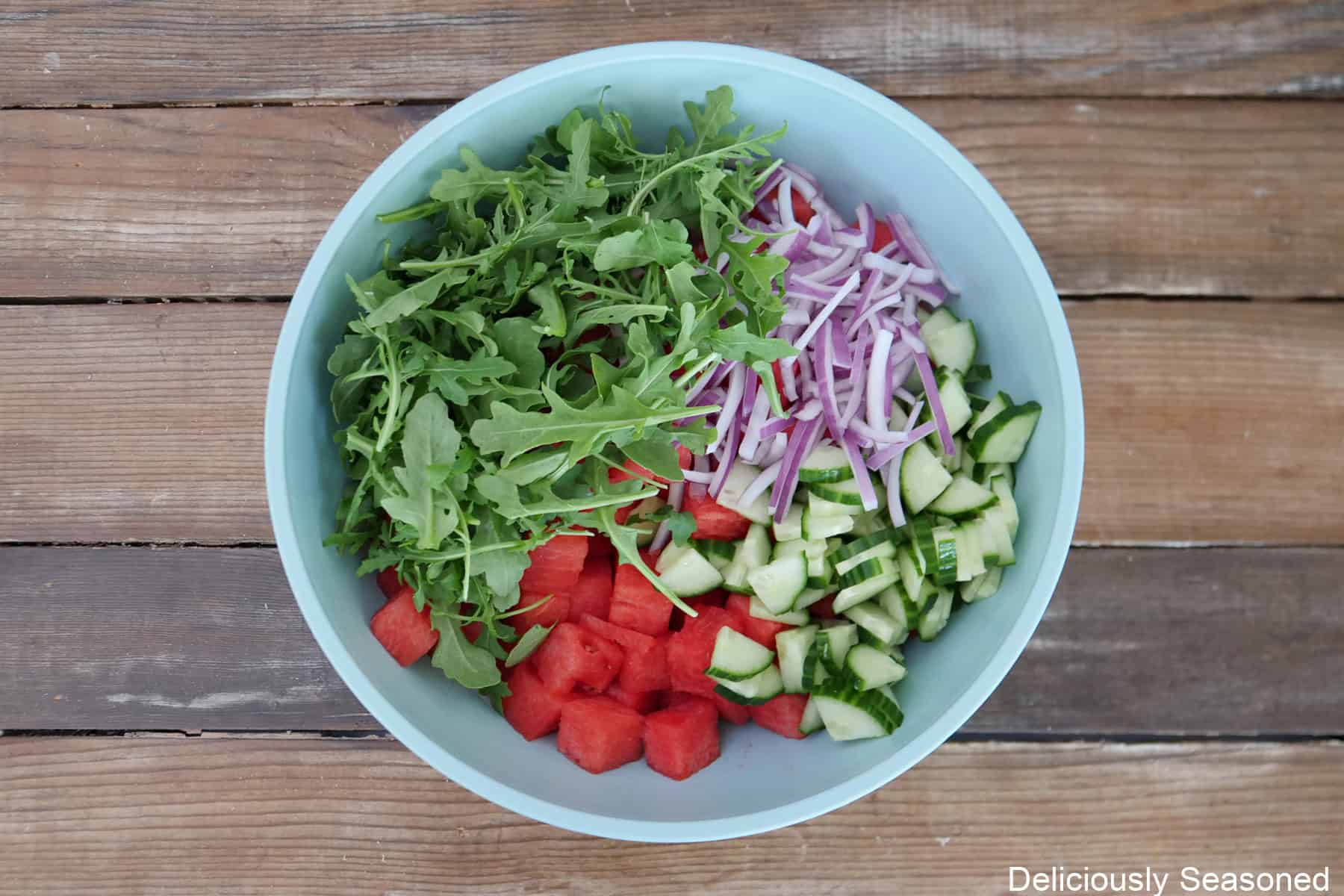 A wood surface with a light blue bowl filled with chopped watermelon, red onions, cucumbers, and arugula in it.