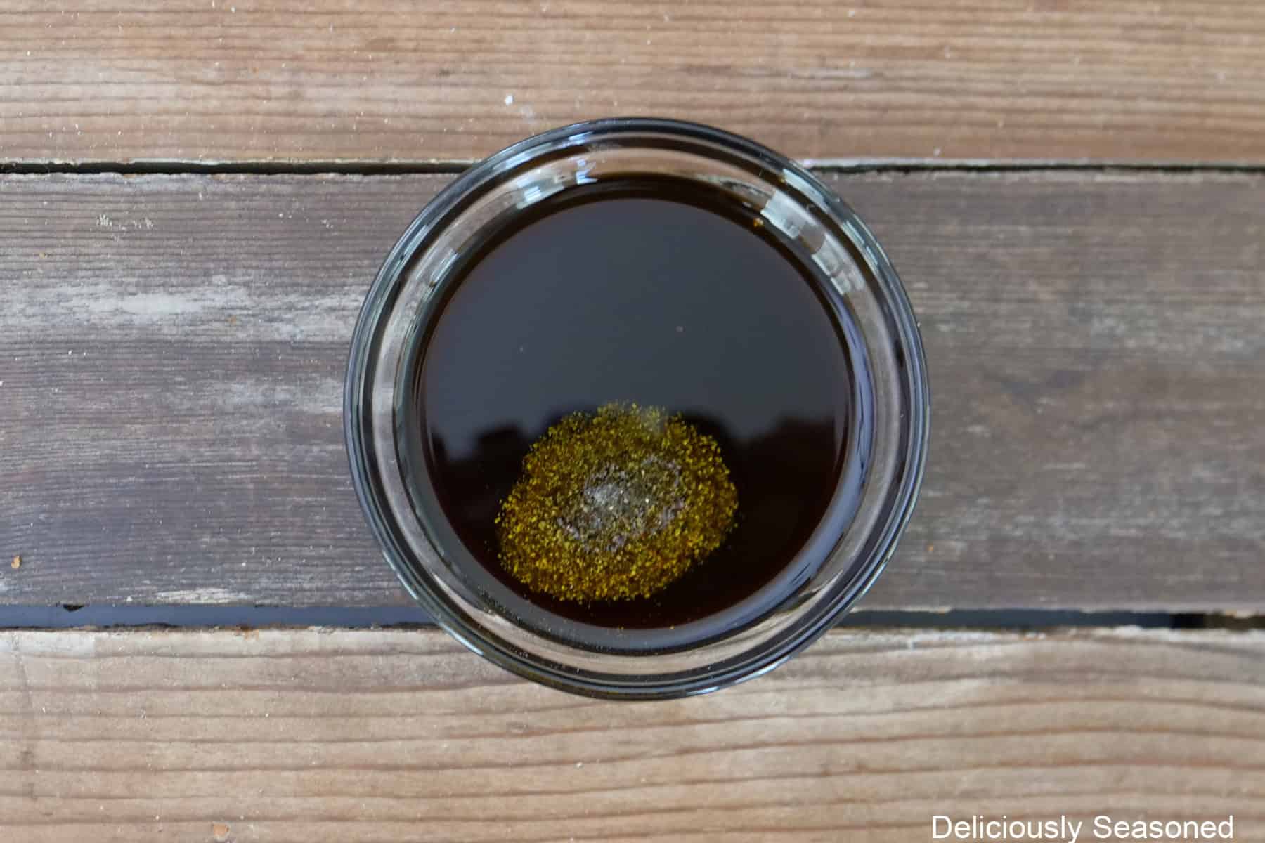 A wood surface with a small glass bowl filled with the balsamic dressing ingredients in it.