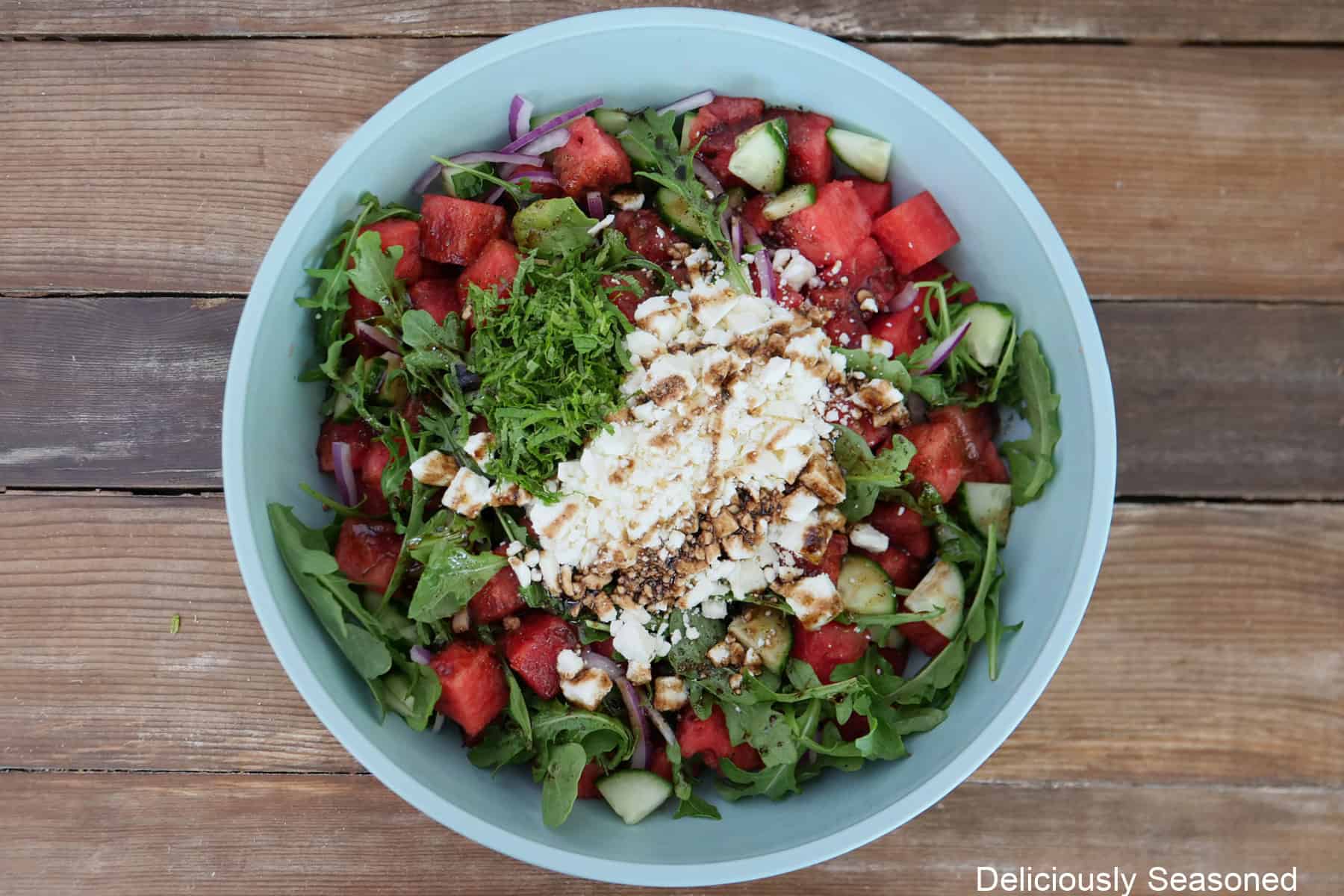 A light blue bowl on a wood surface filled with all the ingredients of a watermelon feta salad in it before being all tossed together.