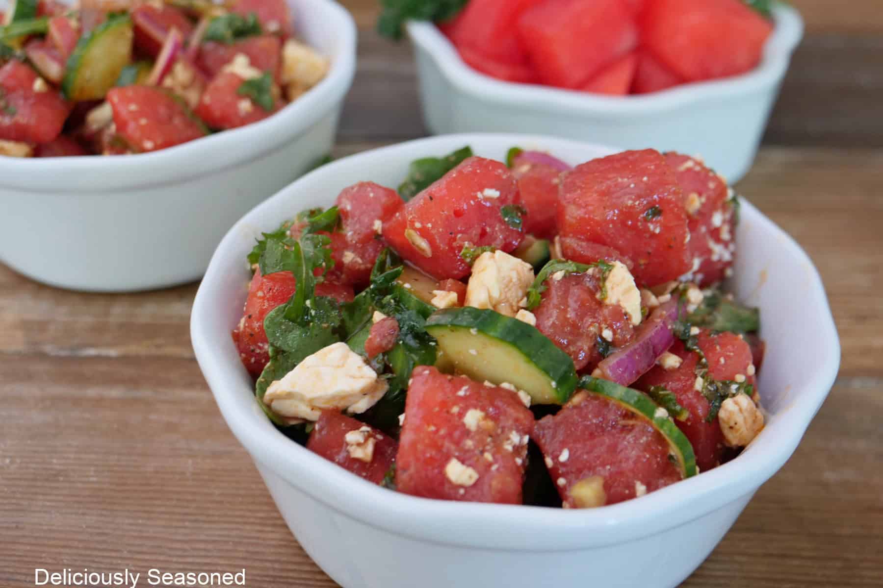 A wood surface with three white bowls, two filled with a serving of watermelon feta salad and one filled with cubed watermelon.