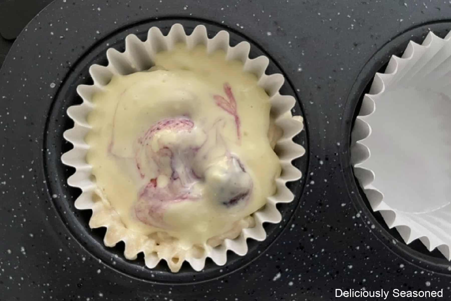 A black muffin tin with one paper liner filled with blueberry cream cheese muffin mixture added and cream cheese mixture added to the top.