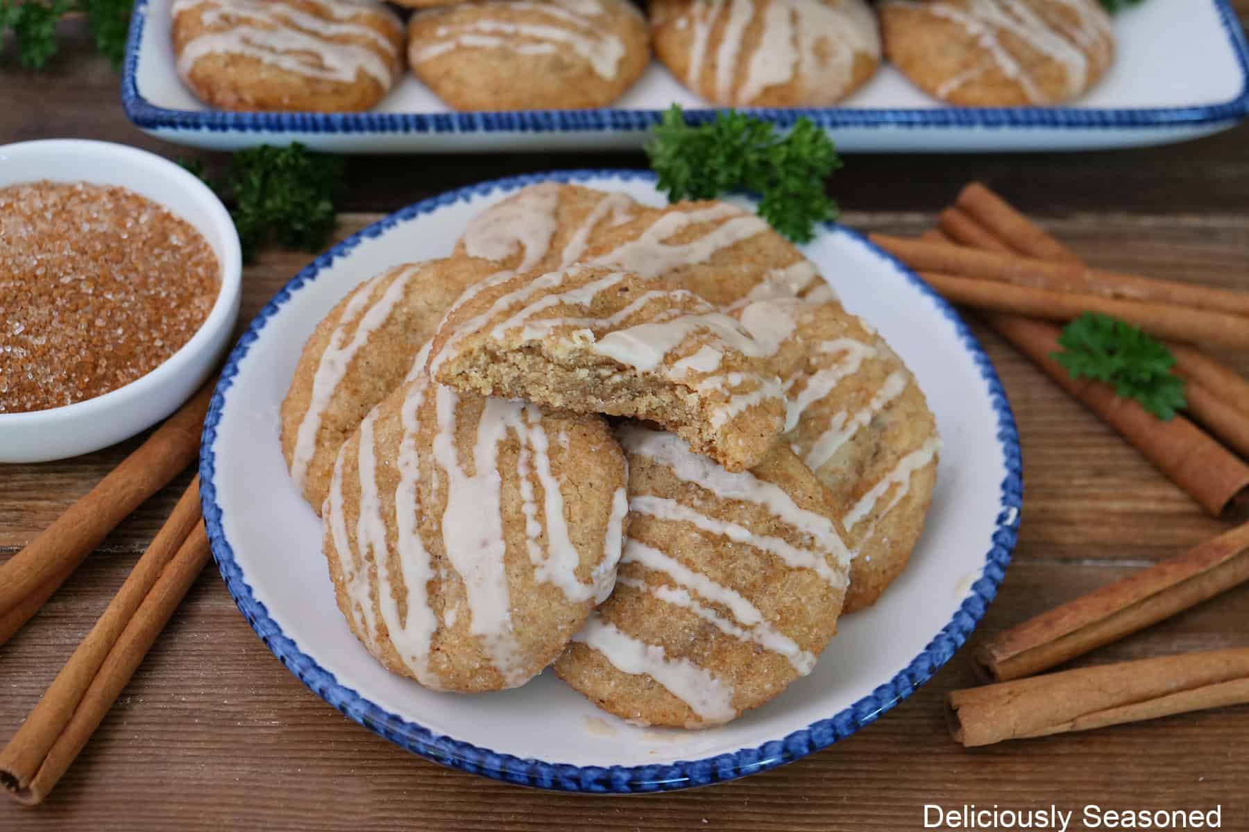 A wood surface with a round white plate with blue trim with five snickerdoodle cookies on it with more in the background, and cinnamon sticks placed beside the plate.