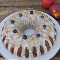 A peach blueberry bundt cake on a white plate with peaches in the background.