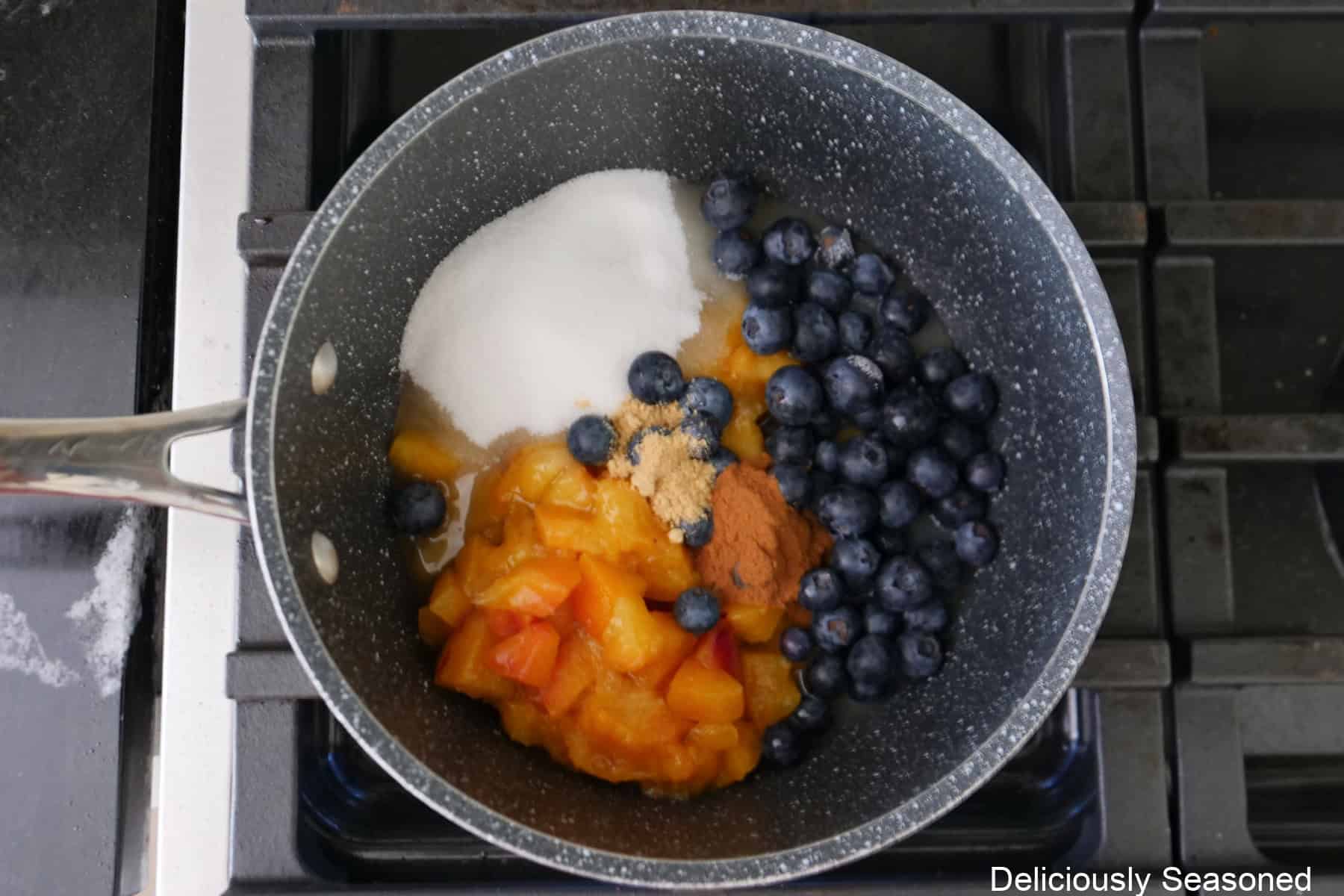 A saucepan with peaches, blueberries, sugar, lemon juice, vanilla, cinnamon and ginger in it before being mixed together.