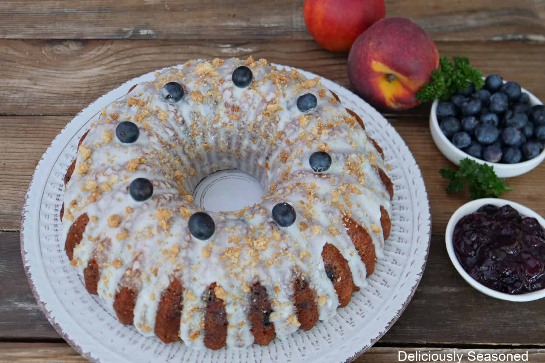 A wood surface with a white plate with a peach blueberry Bundt cake on it that has been frosted with icing and topped with blueberries and streusel, and peaches and blueberries in the background.
