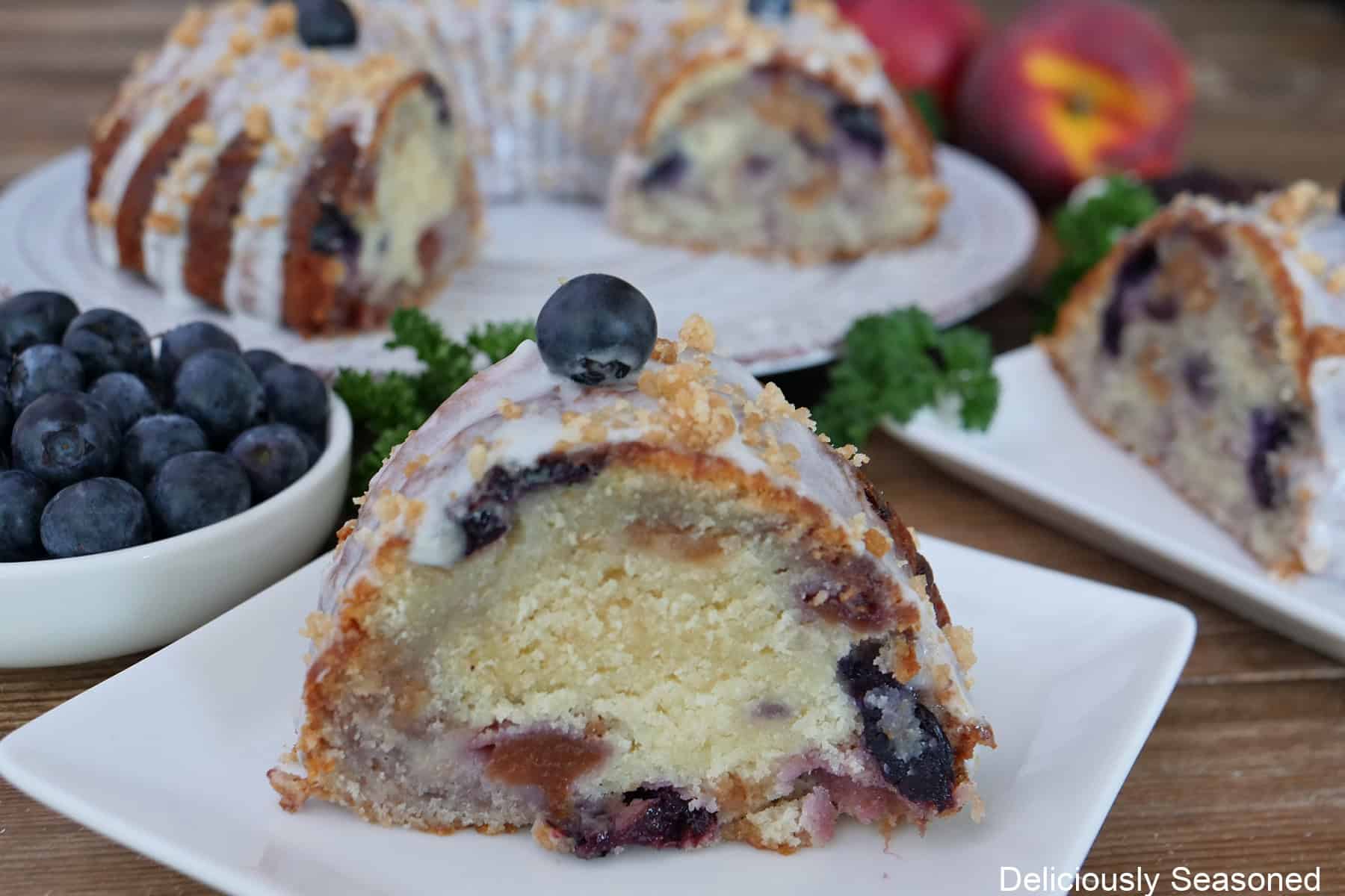 A white plate with a slice of peach blueberry bundt cake on it with the cake on a white plate in the background.