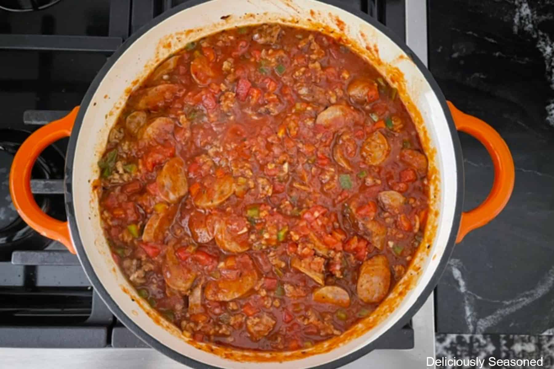 A frying pan on the stove with a tomato base sauce with Italian sausage, onions, peppers, and sliced sausage in it.