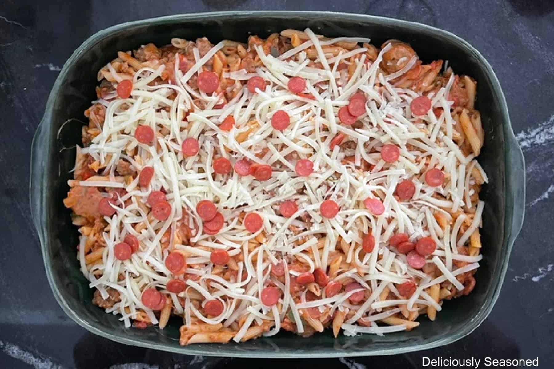 A black countertop with a 9 by 13 green baking dish with Italian sausage pizza pasta in it before being baked.