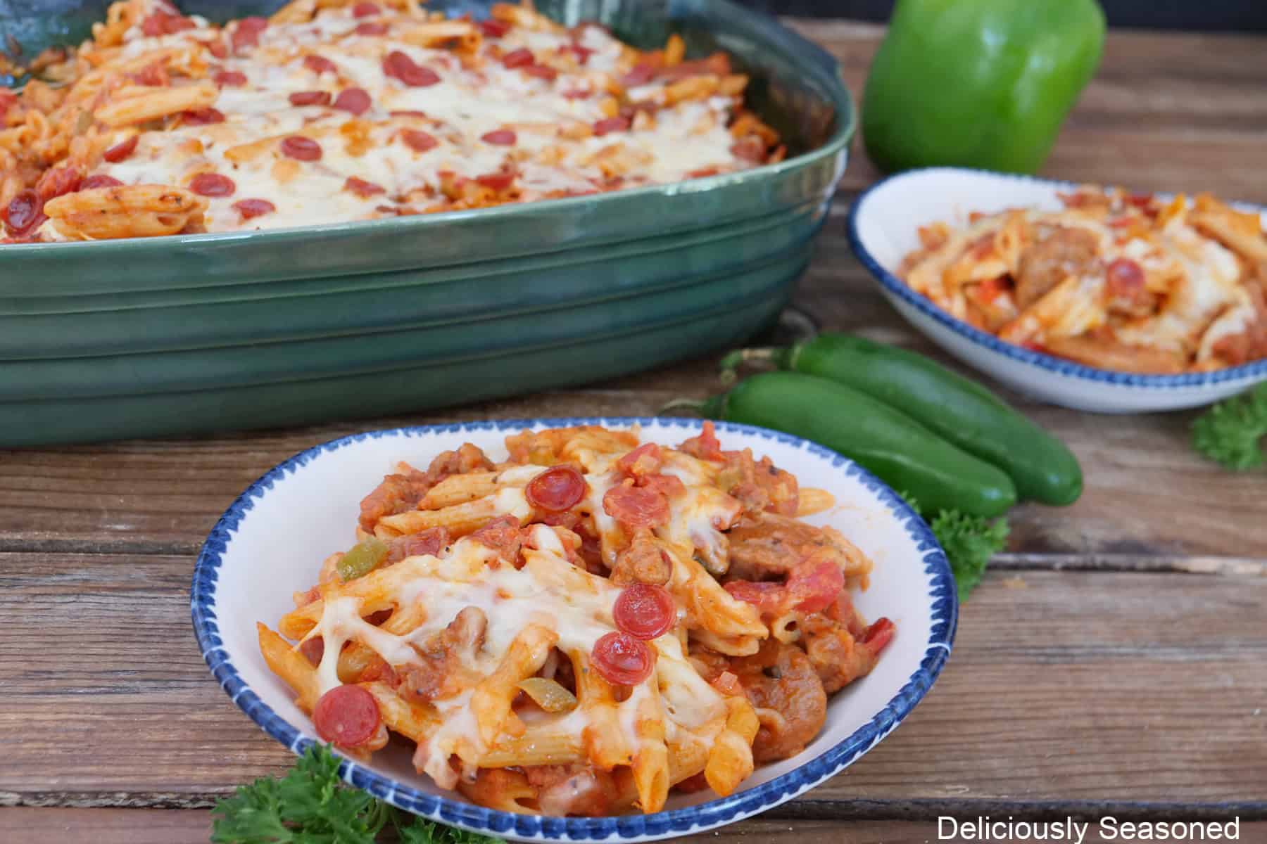A rustic wood surface with tow white bowls with blue trim filled with a serving on pepperoni sausage pizza pasta and a green baking dish filled with the pasta.