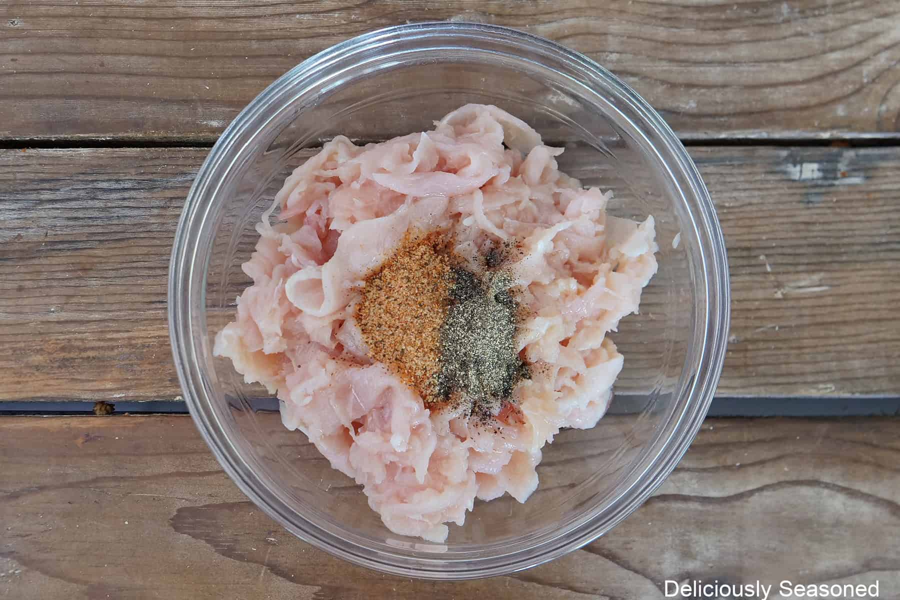 A wood surface with a glass bowl filled with shaved chicken, seasonings, and vegetable oil.