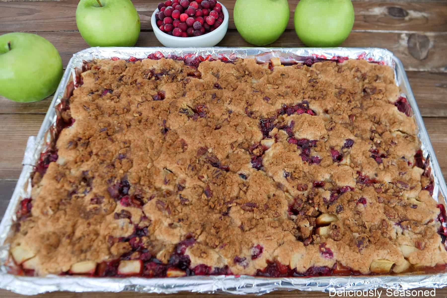 A foil lined baking sheet with cranberry apple cobbler on it with four green apples and a small white bowl of cranberries in the background.