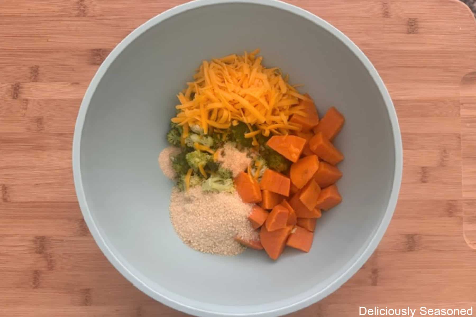 A wood surface with a light blue bowl with all the ingredients for sweet potato broccoli cheddar fritters in it before being mixed together.