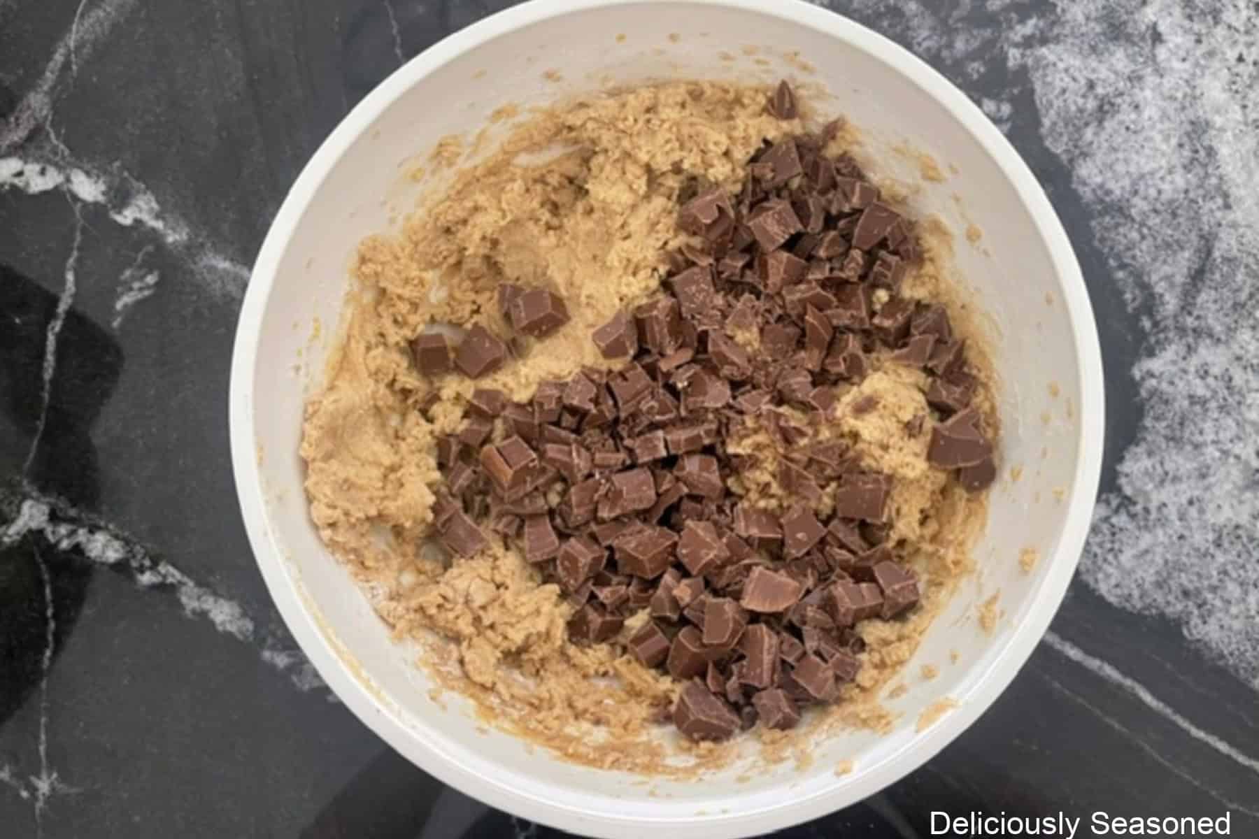 A white bowl with brown butter cookie batter with chocolate bar chopped up and added to the batter.