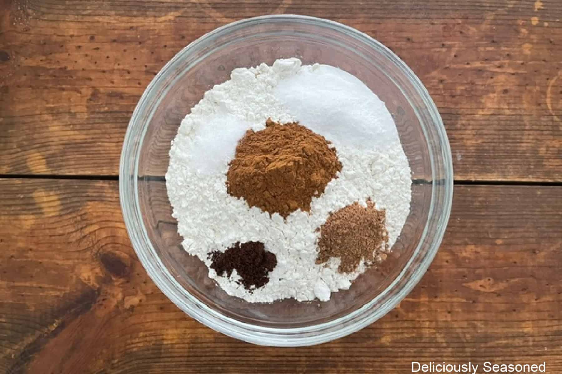 A wood surface with a glass bowl filled with flour, baking soda, salt, cinnamon, nutmeg, and cloves.