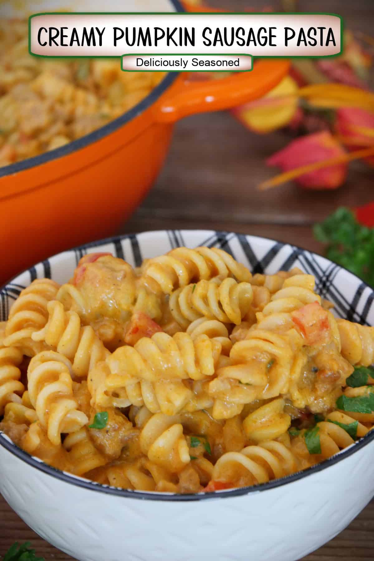 A white bowl with black filled with creamy pumpkin sausage pasta, and fall foliage in the background, and a title overlay at the top.