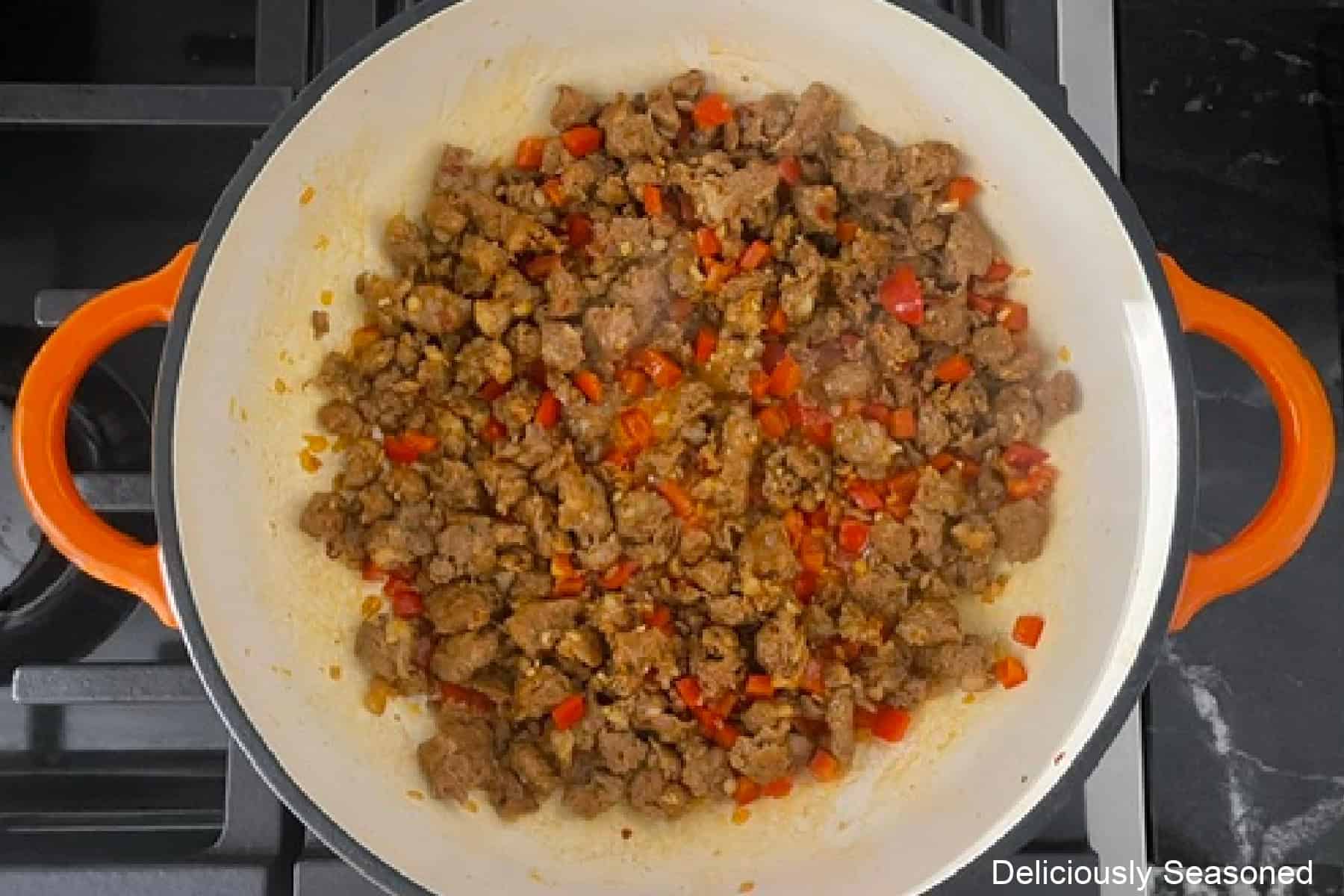 An orange with white cast iron pan with cooked sausage, onions, and red bell pepper in it.