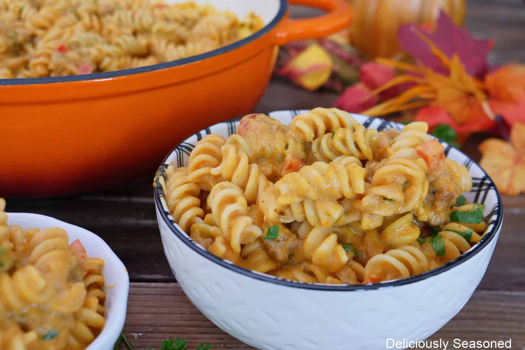 A wood surface with a white bowl filled with a serving of creamy pumpkin sausage pasta with a cast iron pan and fall foliage in the background.