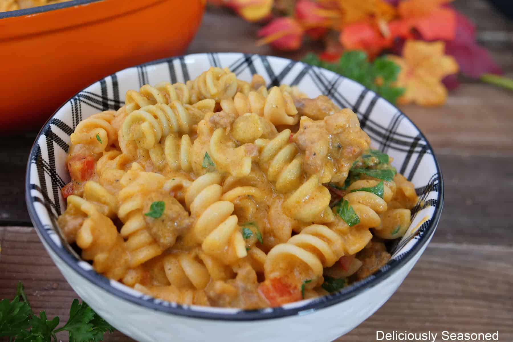 A white bowl with black on the inside filled with a serving of creamy pumpkin sausage pasta, and fall foliage in the background.