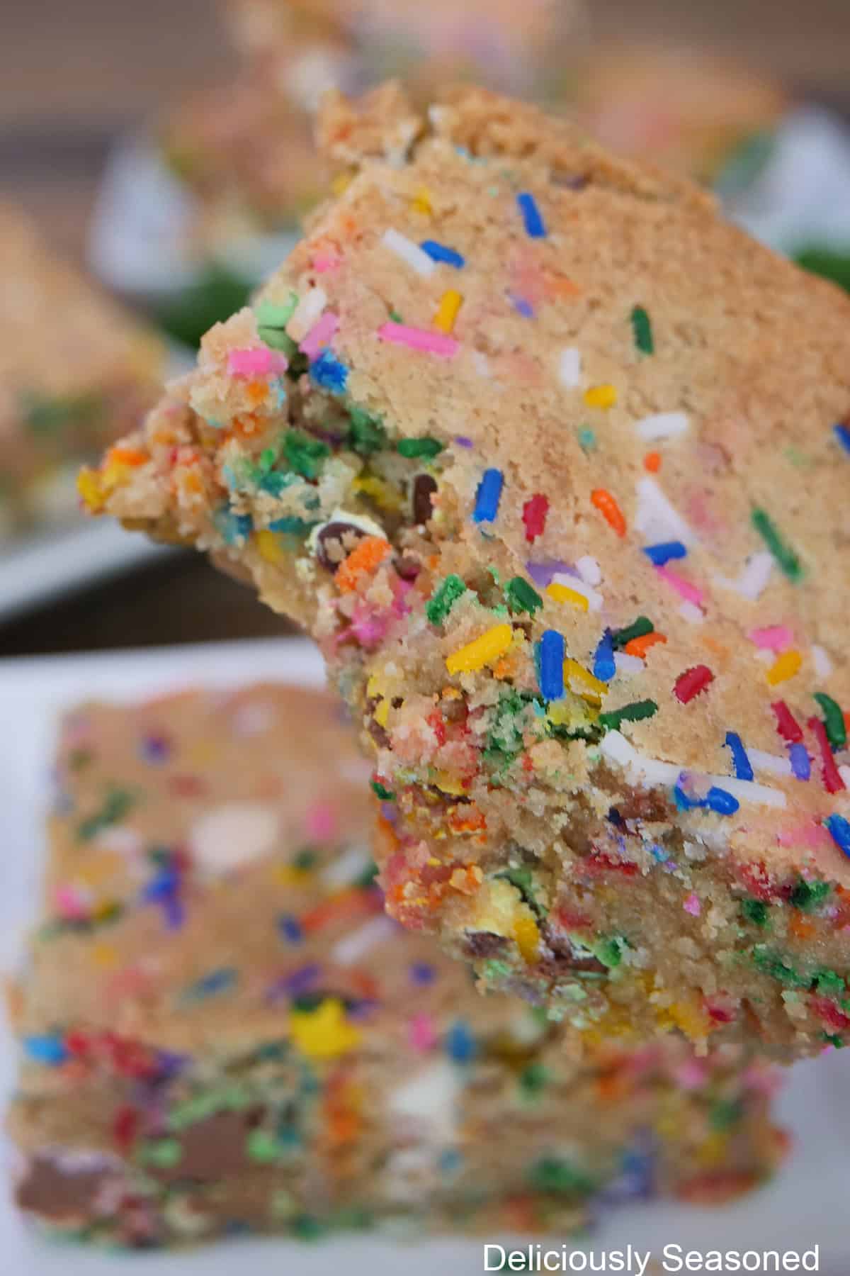 A funfetti blondie with a bite taken out of it held above another blondie on a white plate below.
