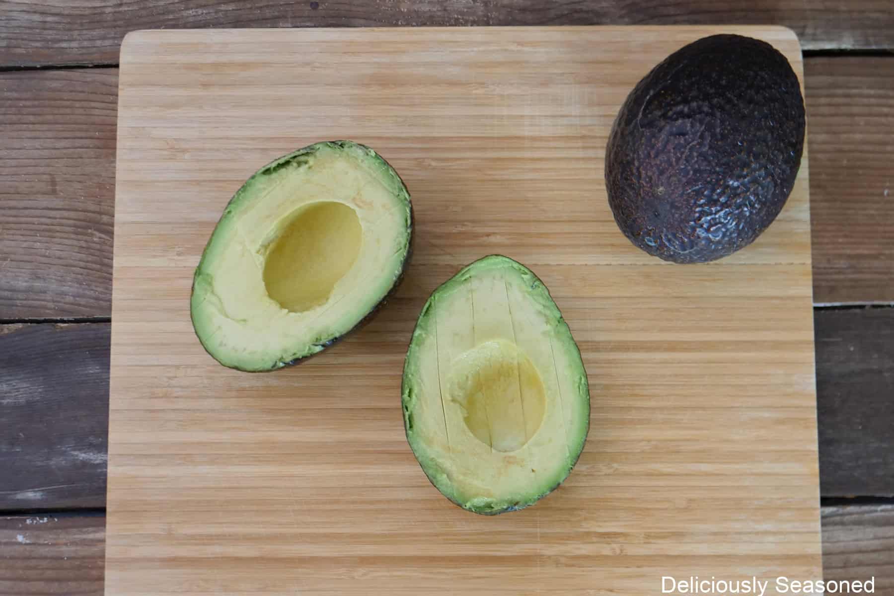A wood cutting board with two avocados on it, one cut in half, seed removed, and the flesh sliced while still in the peel, and a whole avocado.