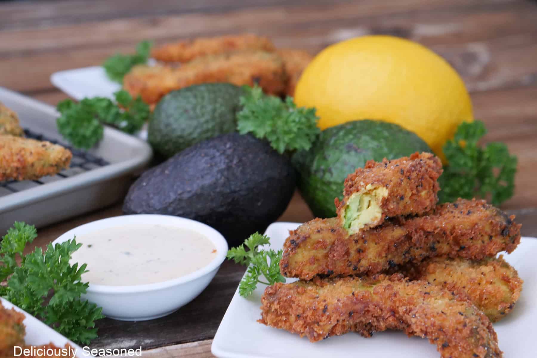 A white plate with lemon pepper avocado fries on it with a small bowl of ranch dressing, a lemon and more avocados in the background.