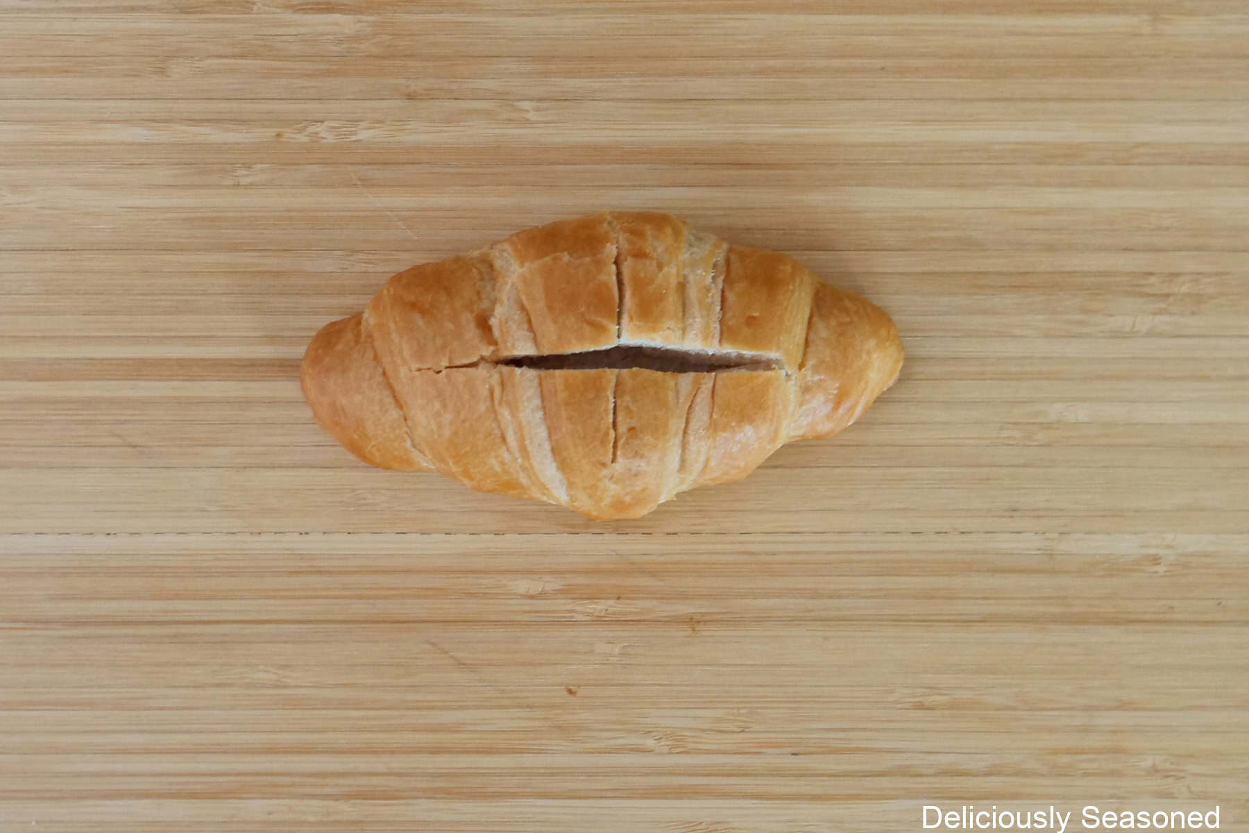 A wood surface with a mini croissant placed on it with a cut down the top of the croissant.