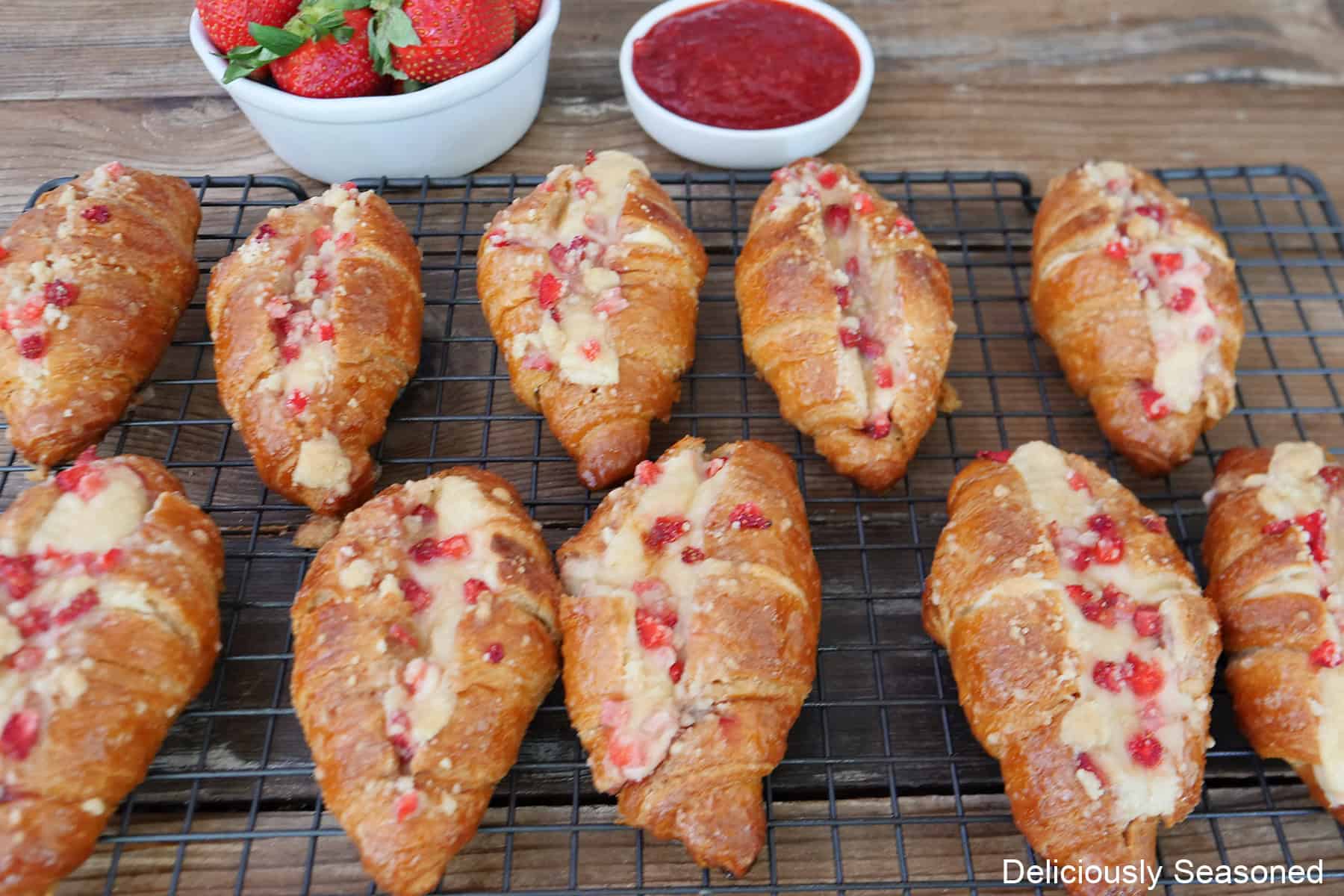A wire rack with ten baked mini strawberry cheesecake croissants on it with a small bowl of strawberry filling and fresh strawberries in the background.