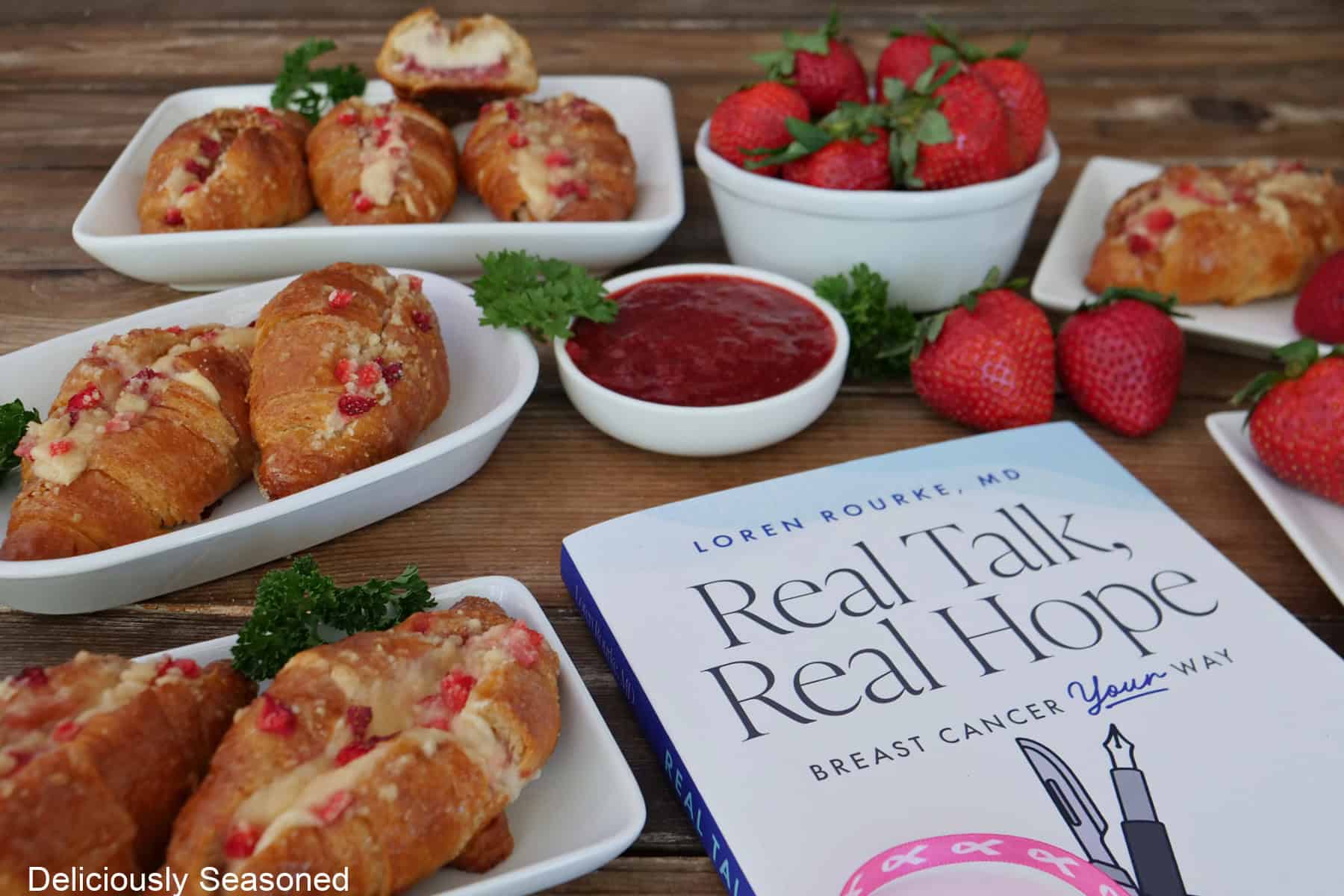 A wood surface with white plates with mini strawberry cheesecake croissants on them and a copy of Dr. Loren Rourke's book, Real Talk, Real Hope next to the croissants.