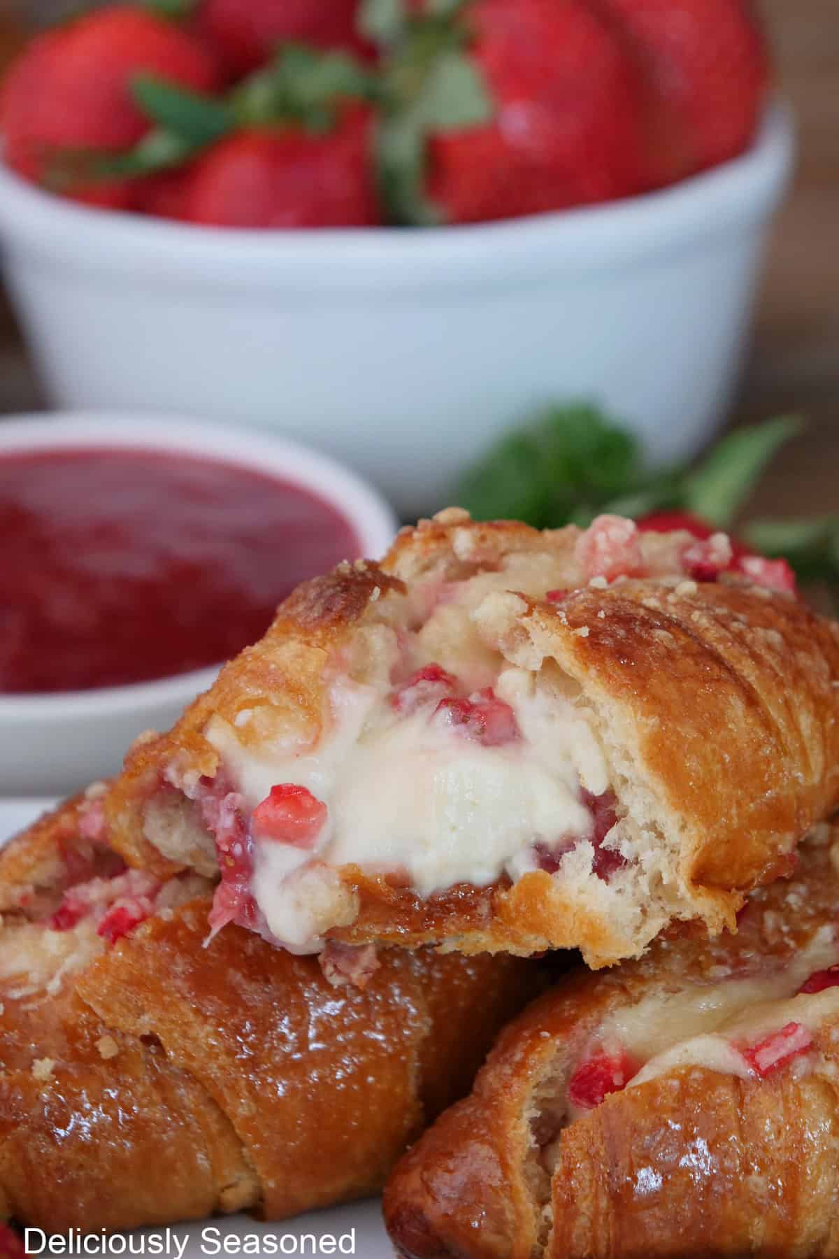 Three mini strawberry cheesecake croissants stacked on a white plate with a bite taken out of the one on top, and fresh strawberries and strawberry filling in the background.