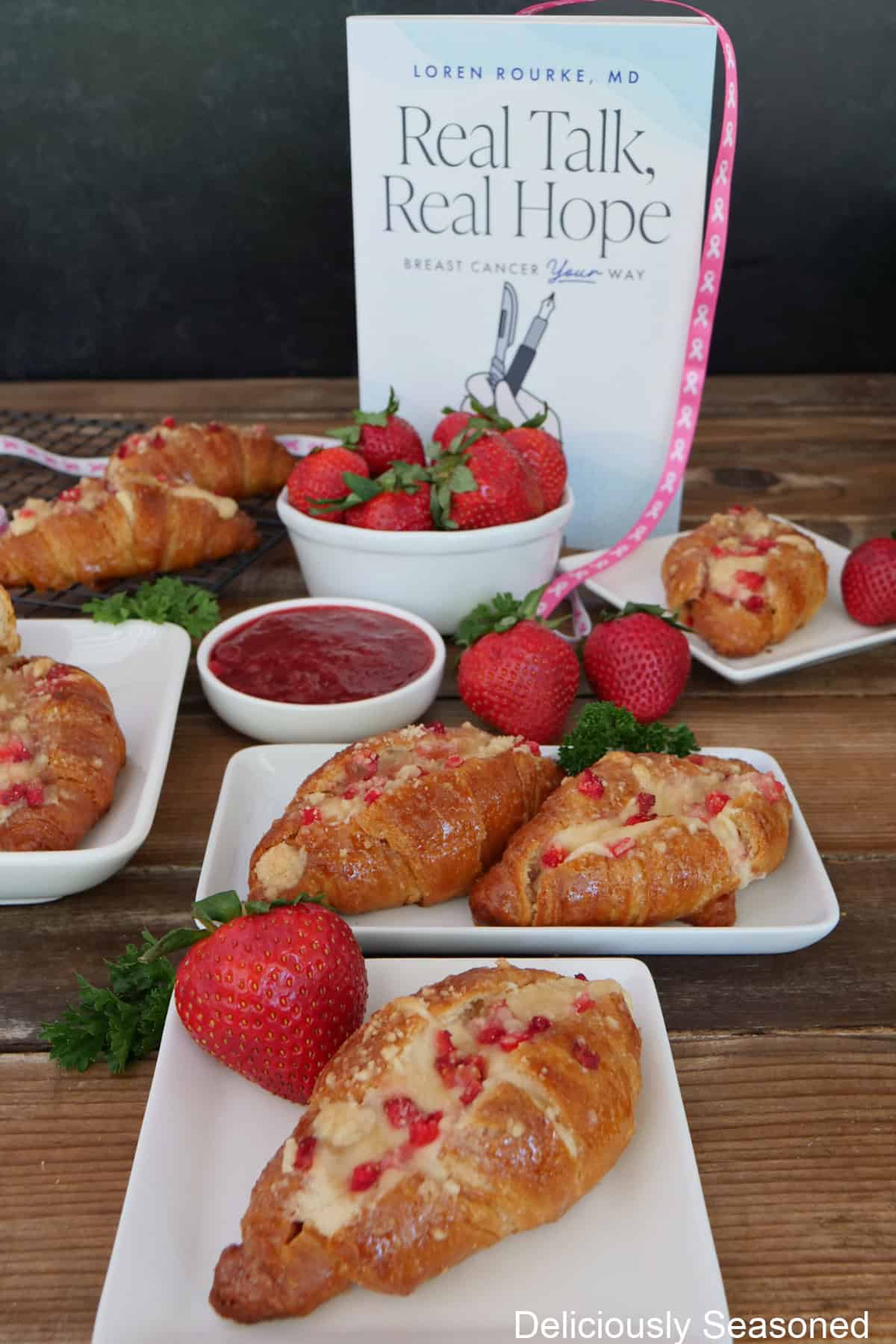 A wood surface with mini strawberry cheesecake croissants on white plates, and a copy of Real Talk, Real Hope by Dr. Loren Rourke.