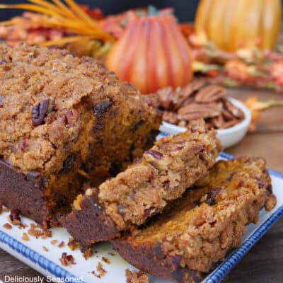 A white plate with blue trim with a pumpkin chocolate chip loaf on it with two slices cut, and fall foliage in the background.