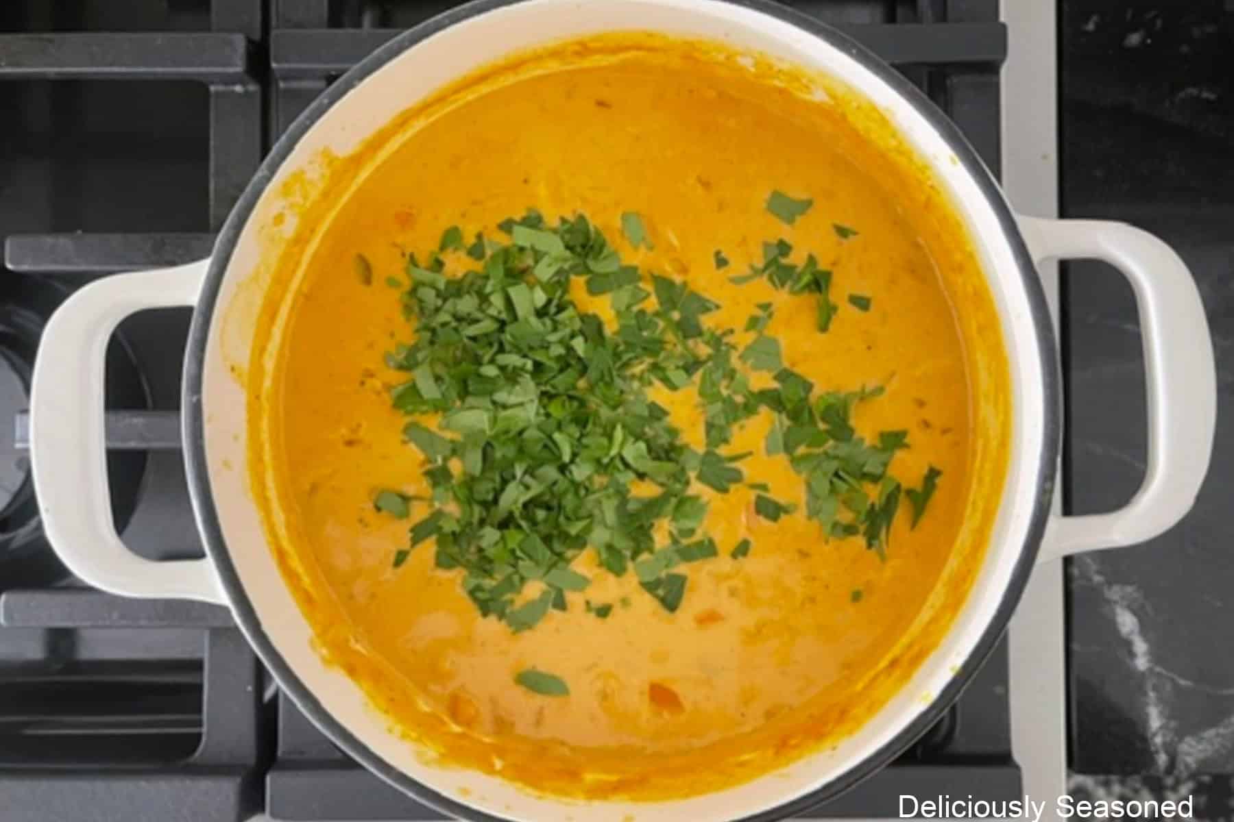 A cast iron white pot with creamy pumpkin gnocchi soup in it with chopped parsley added before being stirred together.