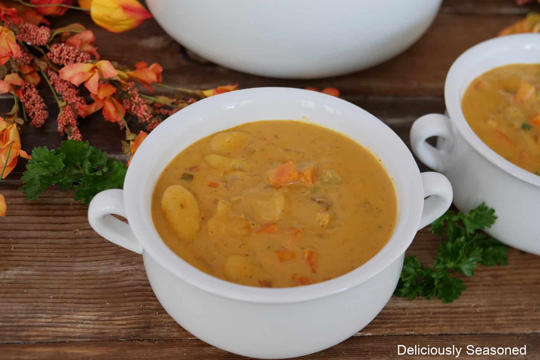 A wood surface with two white soup bowls filled with pumpkin gnocchi soup, and fall foliage in the background.