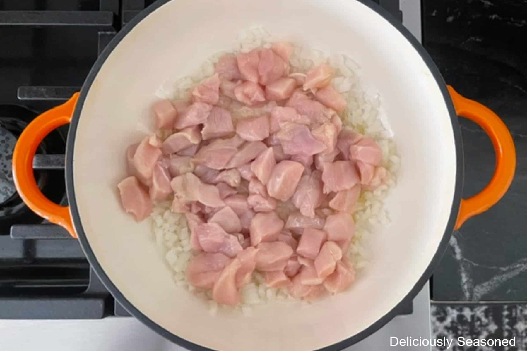 A cast iron pan with diced onions and chunks of chicken pieces in it cooking.