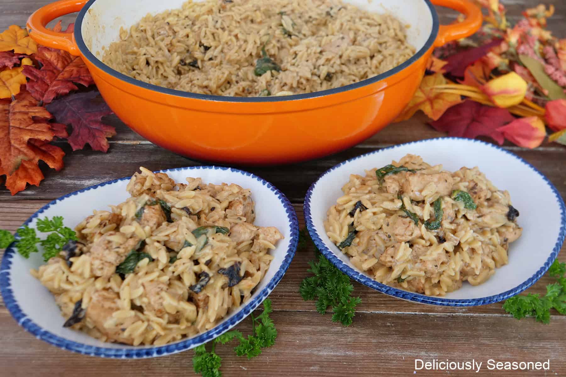 An orange cast iron pan and two white bowls with blue trim with Tuscan chicken orzo in each of them.