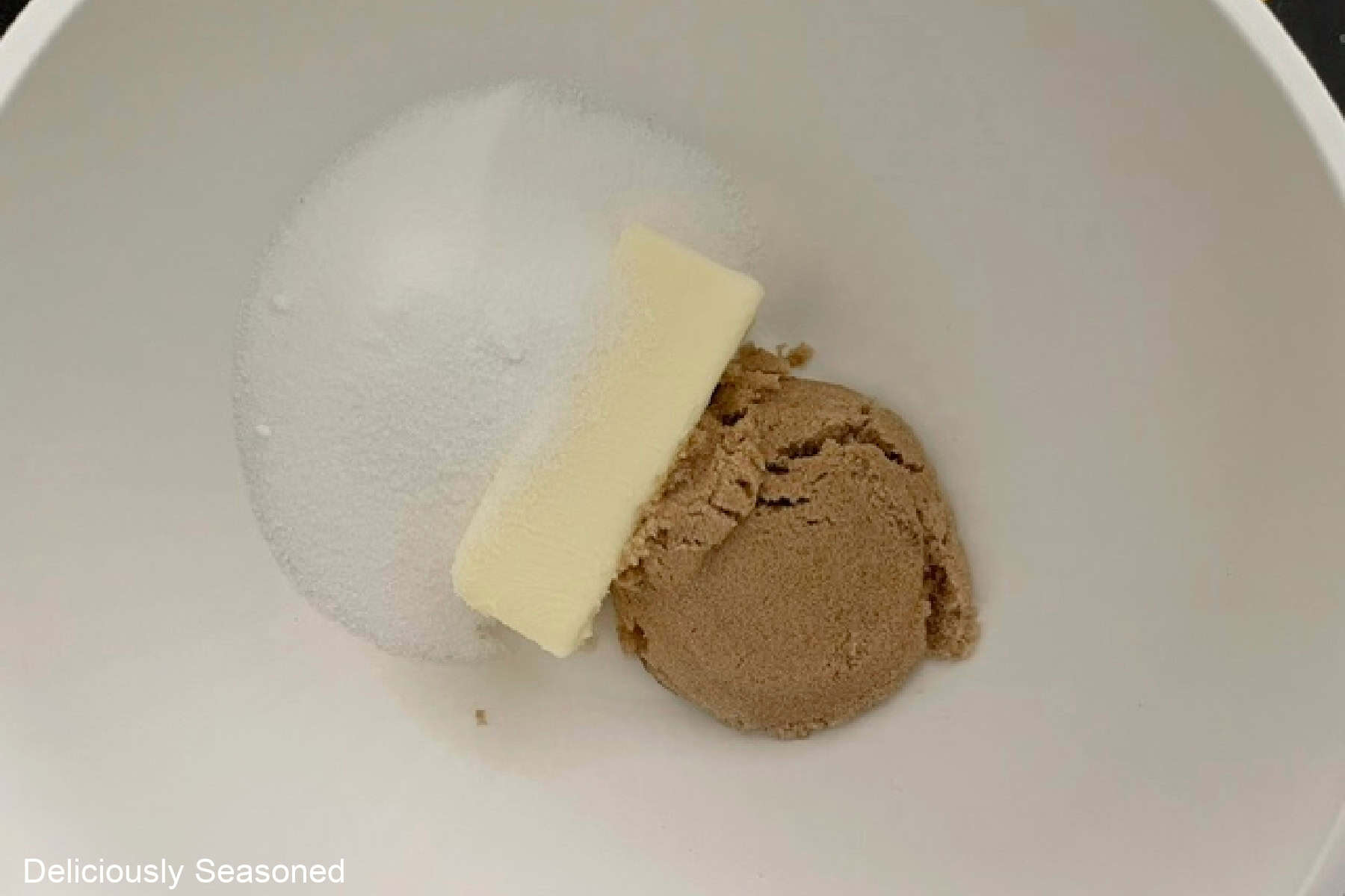 A white mixing bowl with a stick of butter, sugar, and brown sugar in it.