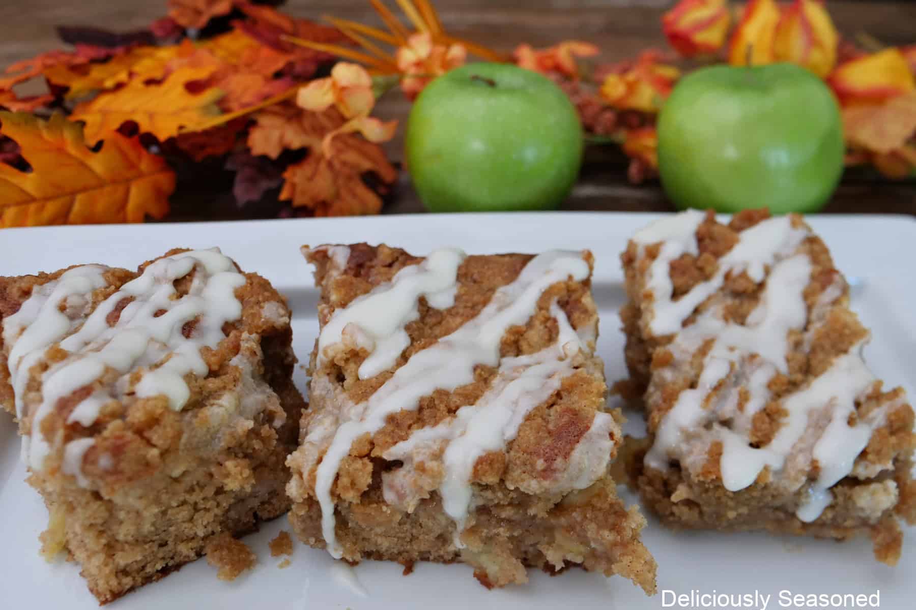 A white plate with three servings of apple cider coffee cake with maple icing, two green apples and fall foliage in the background.
