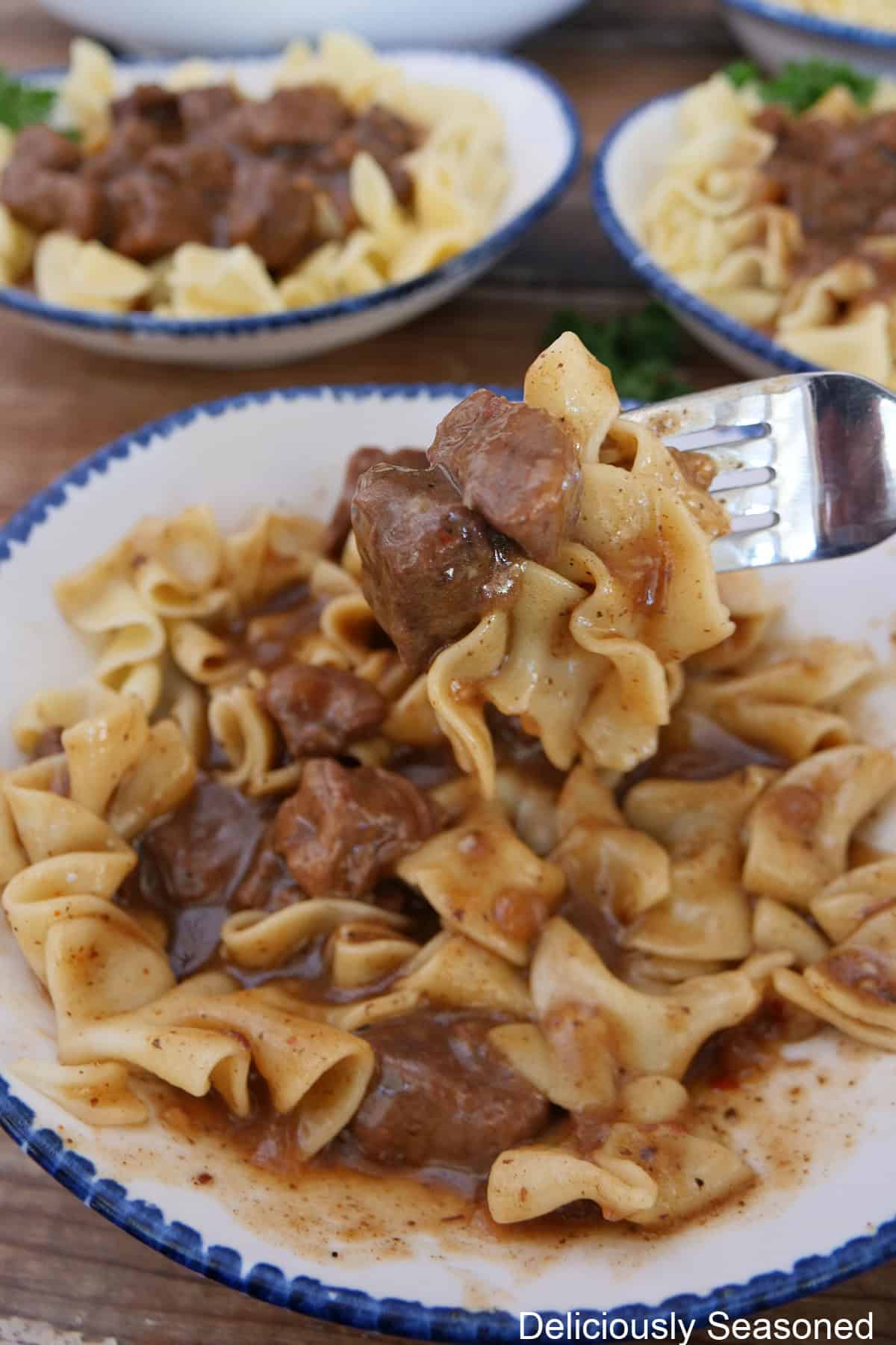 A white bowl with blue trim with a serving of beef and noodles and a bite on a fork held slightly above the bowl.
